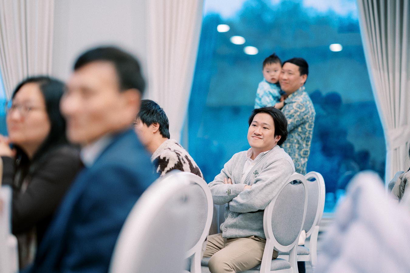 A group of people attends a formal indoor event, with a focus on a smiling man in a gray sweater seated in the center. A man holding a child is seen in the background, standing near a large window with blue light.