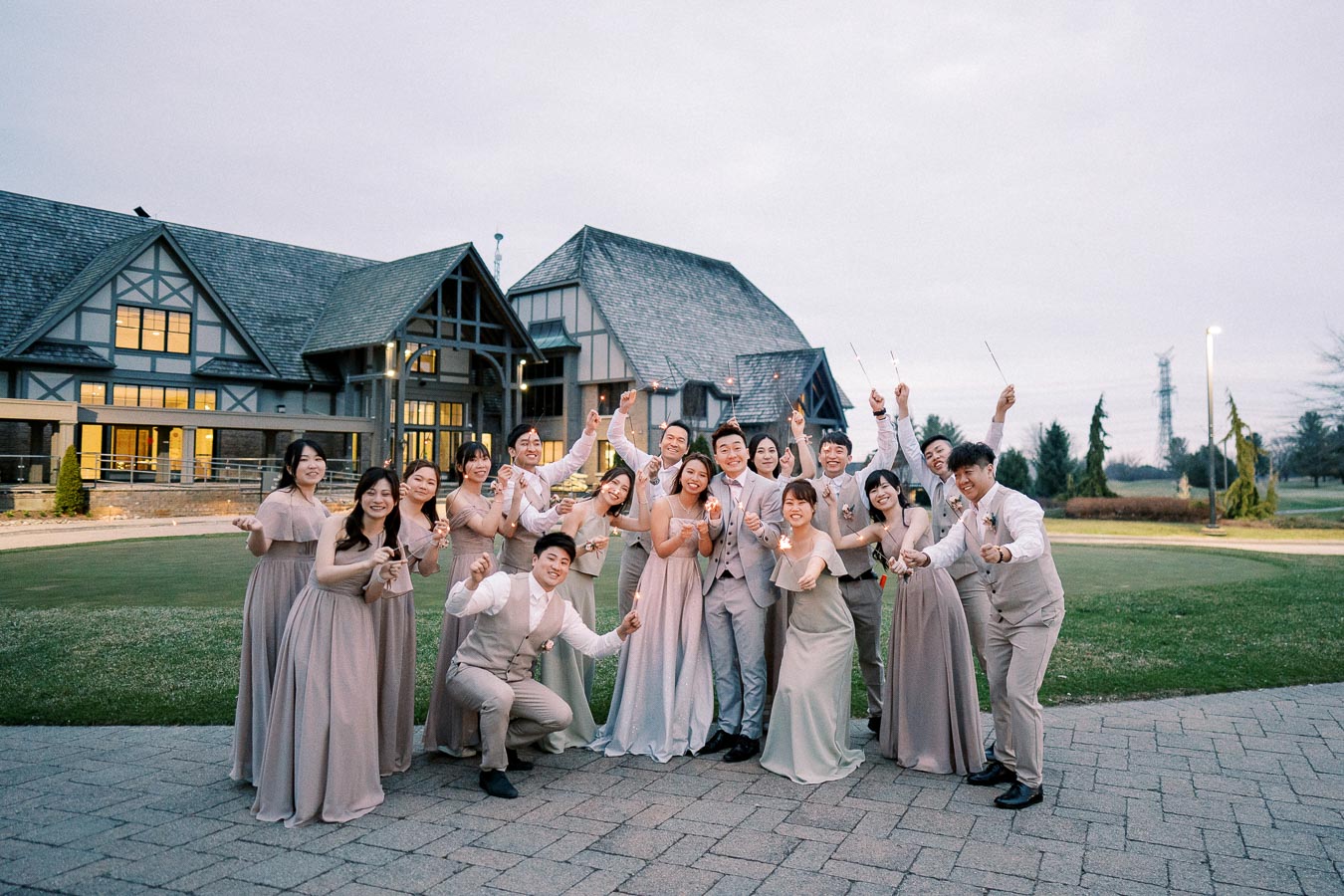 A joyful wedding party posing with sparklers in front of a large, elegant mansion at dusk, featuring bridesmaids in taupe dresses and groomsmen in beige suits, celebrating outdoors on a well-manicured lawn.