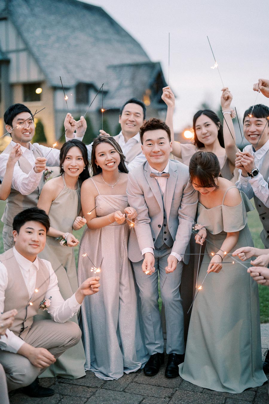 A joyful wedding party gathered with sparklers, featuring a bride in an elegant gown and a groom in a light suit, surrounded by bridesmaids and groomsmen in matching attire, celebrating outdoors near a rustic building.