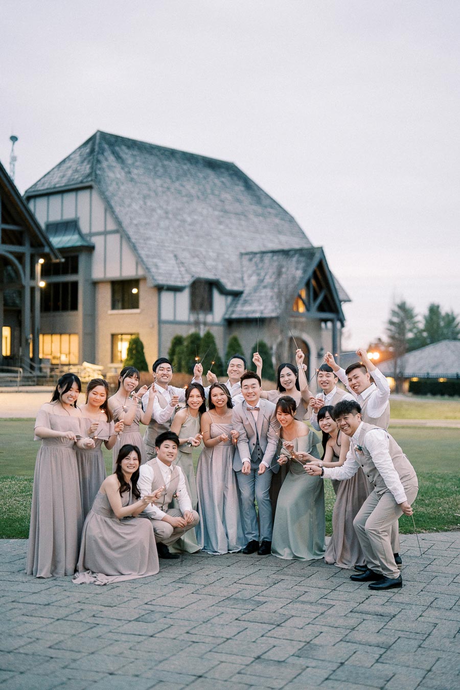 A large wedding party, dressed in light pastel suits and dresses, holding sparklers in front of a rustic, timber-framed building at dusk.
