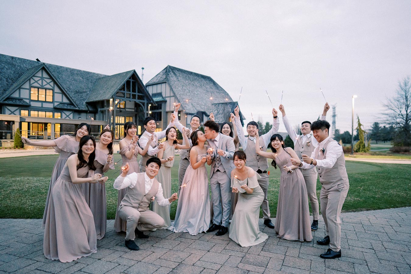 Wedding celebration with a joyful bridal party holding sparklers outside a rustic venue, featuring bridesmaids in elegant dresses and groomsmen in matching suits.