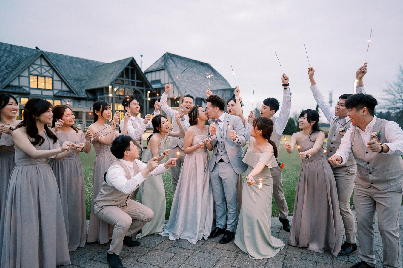 A joyful bridal party celebrating with sparklers outside a rustic venue, featuring a happy couple in formal attire surrounded by enthusiastic friends in matching dresses and suits.