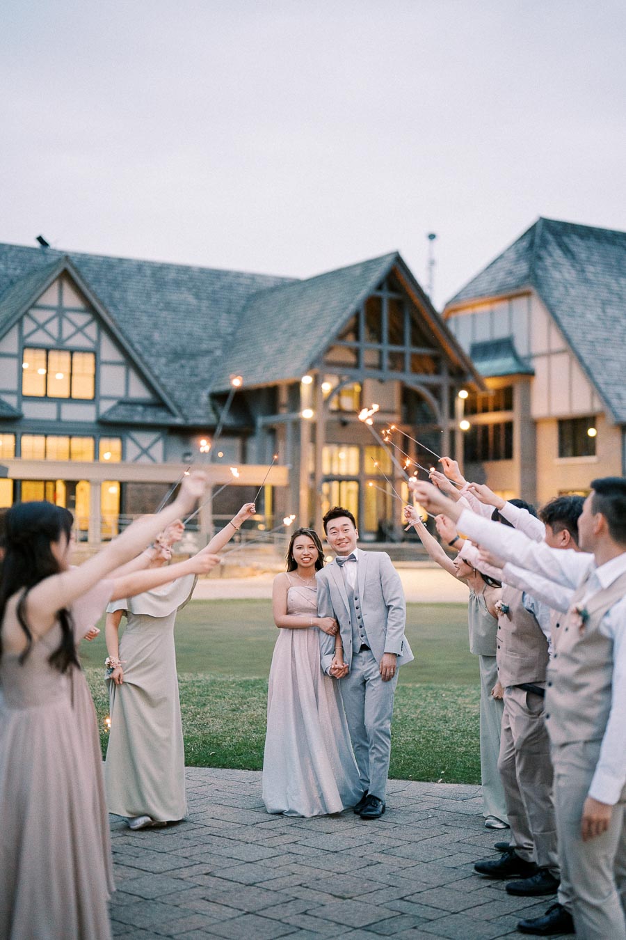 Wedding couple smiling under a sparkler arch, surrounded by guests in elegant attire, outside a beautiful tudor-style building at dusk