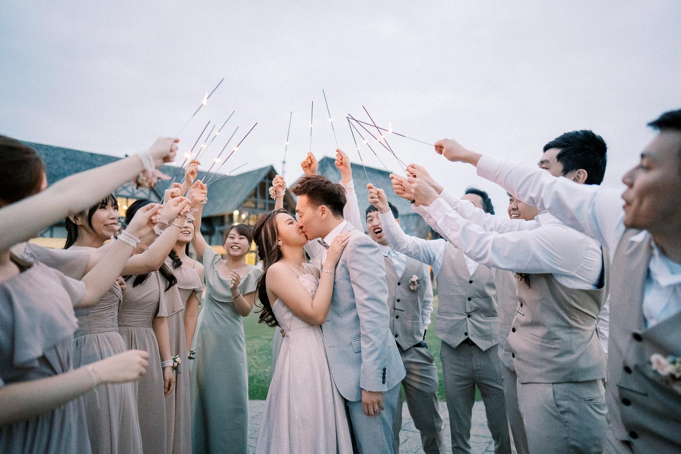A newlywed couple sharing a kiss surrounded by wedding guests holding celebratory sparklers, all dressed in formal attire, with a picturesque outdoor venue in the background.