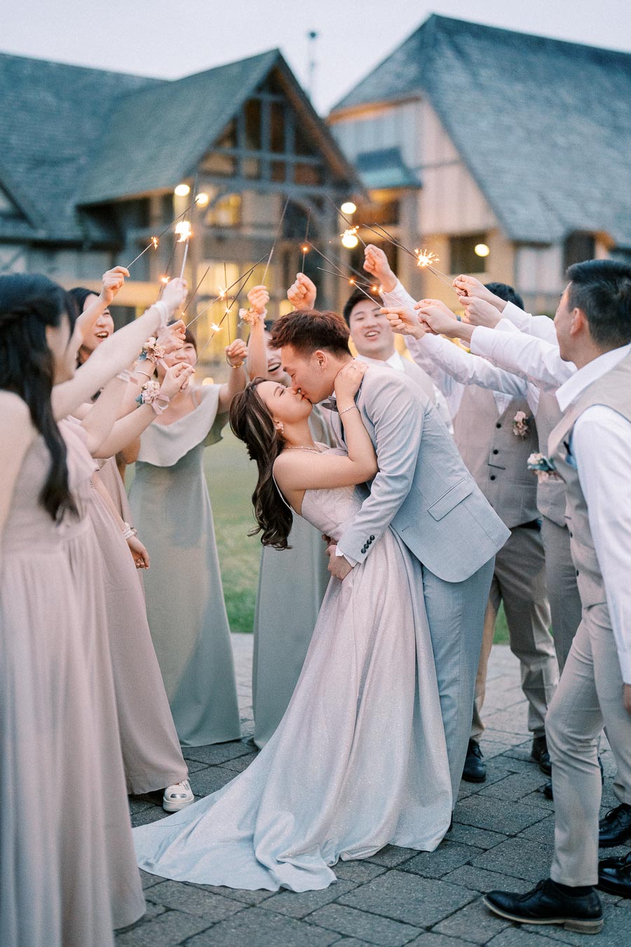 Bride and groom sharing a romantic kiss surrounded by their wedding party holding sparklers, celebrating at an outdoor evening reception.