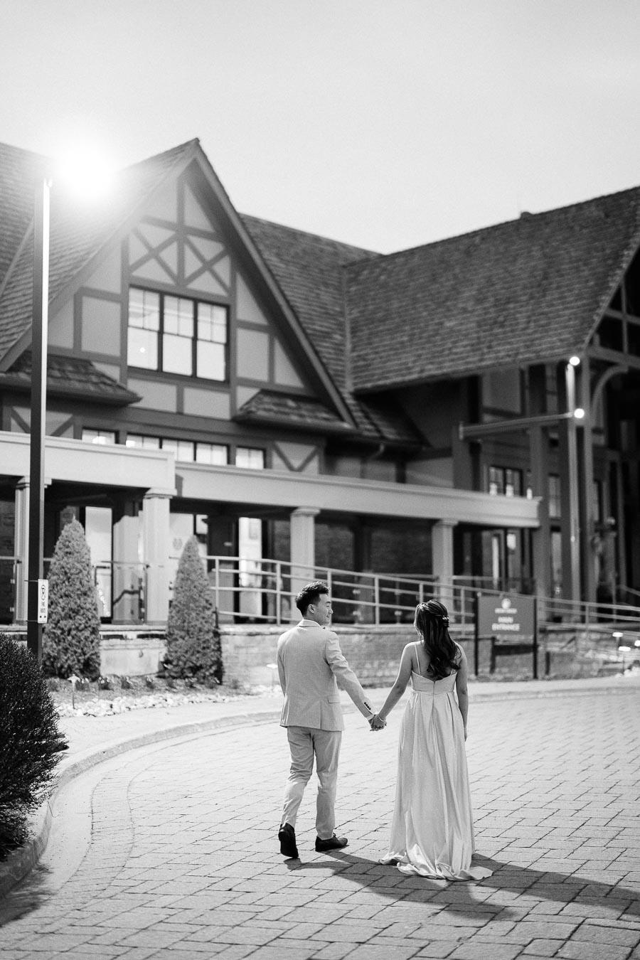 A couple dressed in elegant attire walks hand in hand on a cobblestone path, with a charming Tudor-style building in the background illuminated by streetlights.