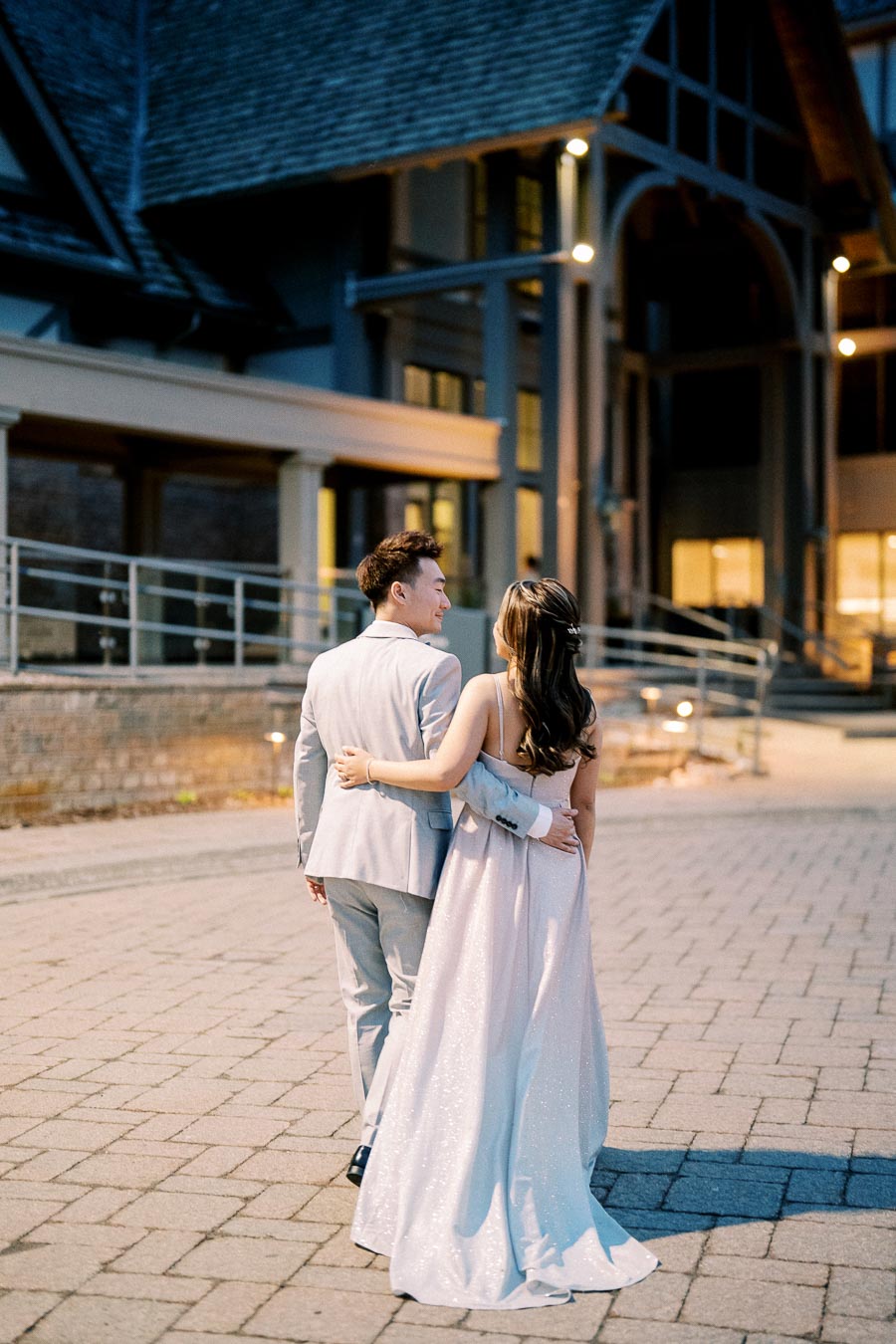 A couple in formal attire, with the woman wearing a glittering gown and the man in a light gray suit, walking hand-in-hand on a cobblestone path towards an elegant building at dusk.