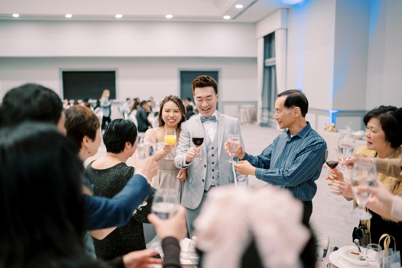 A group of people celebrating at an indoor event, raising glasses in a toast with a joyful atmosphere. The man in a light-colored suit and bow tie stands in the center next to a woman in a light dress. The background features a modern, spacious venue.