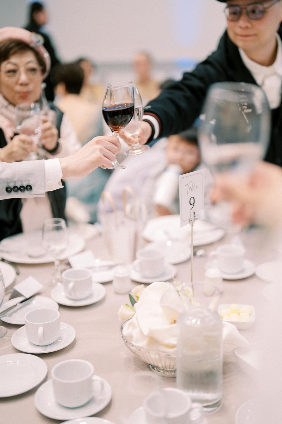 A group of people making a toast with wine glasses at a formal dining table, featuring a table setting with white crockery and a table 9 marker.