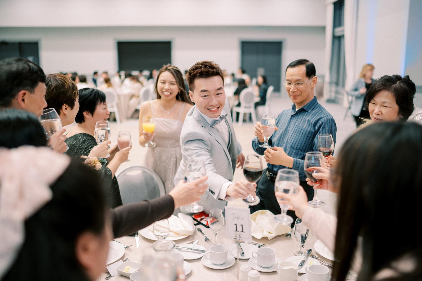 Group of people toasting at a celebratory event, with a well-dressed individual in a light gray suit smiling and leading the toast at a dinner table.