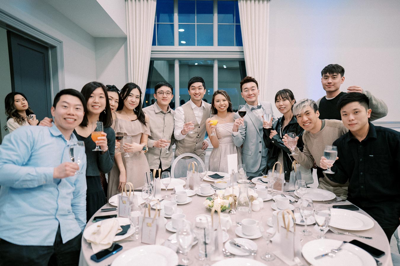 A group of people smiling and holding drinks while gathered around a table set for an event celebration indoors.