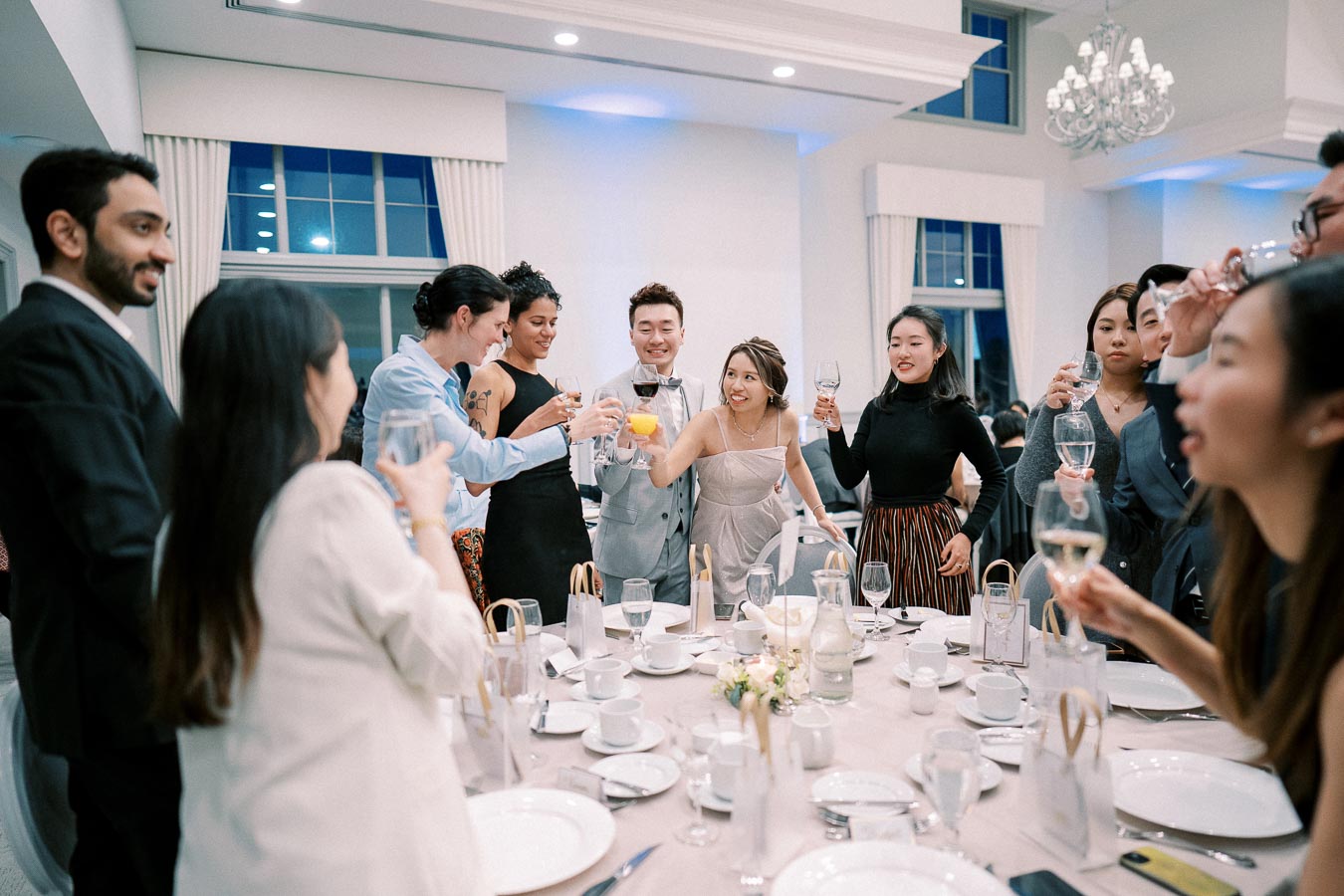 Group of people celebrating at a formal event, raising glasses in a toast around a beautifully set table with white tableware in an elegant room with chandeliers.