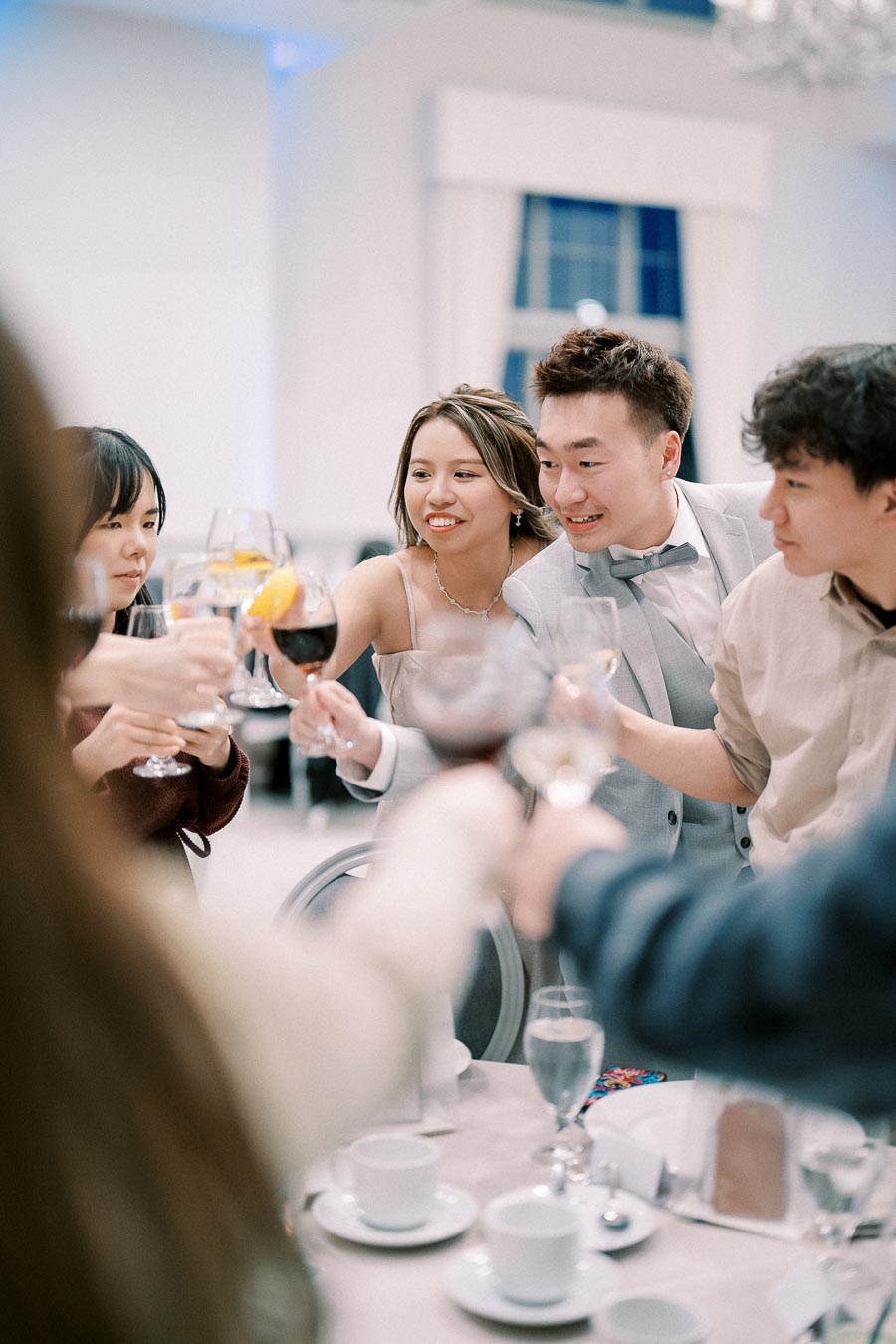 A group of well-dressed young adults clinking glasses in celebration around a table with cups and plates, indicating a festive gathering or party in a bright room.