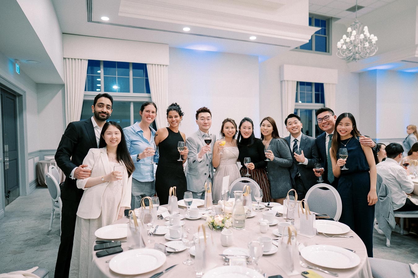 A group of well-dressed individuals smiling and holding drinks at a formal banquet event, gathered around a table set with plates, cutlery, and centerpieces in an elegant venue with chandeliers and large windows.