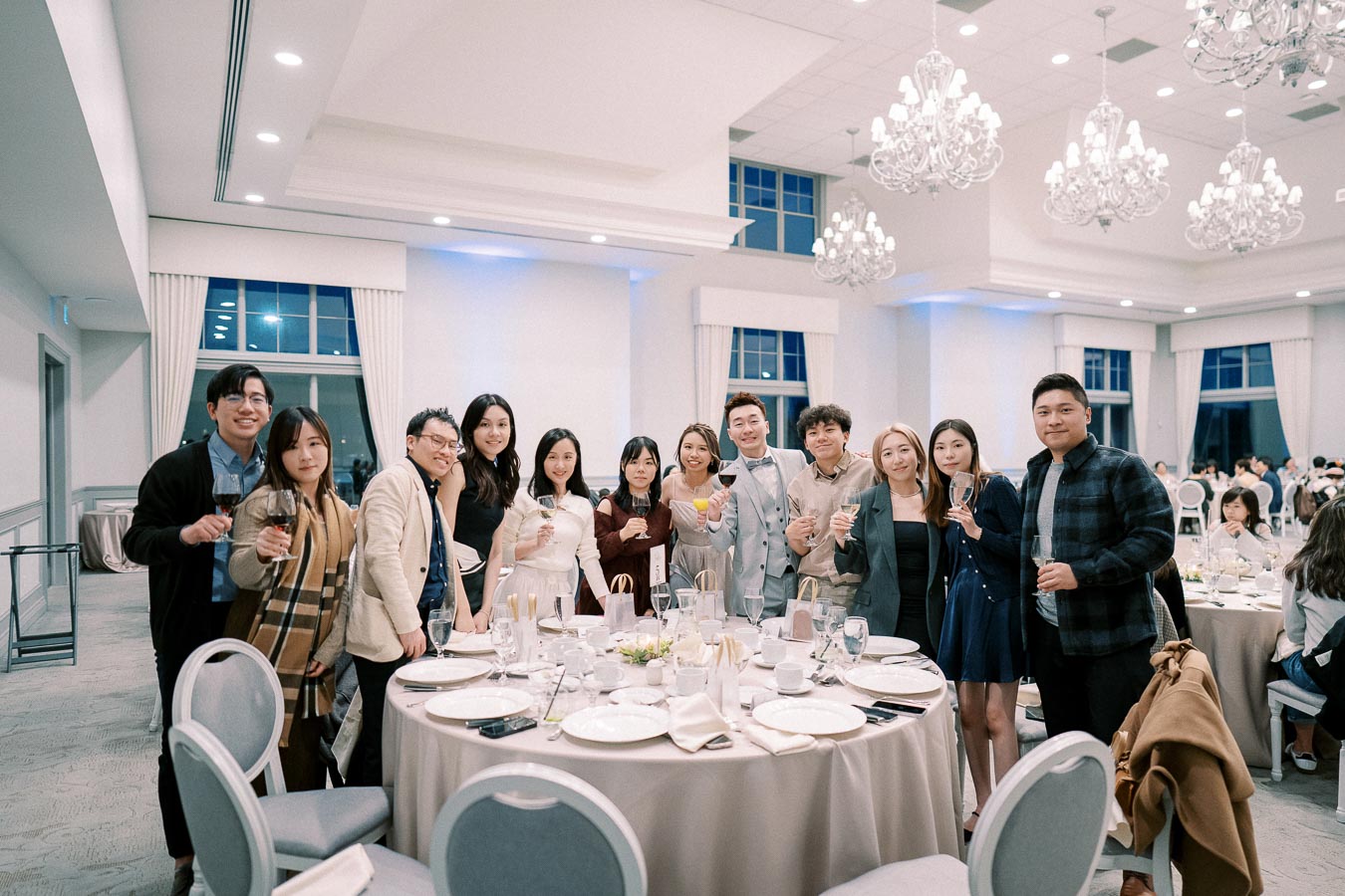 A group of people dressed in formal attire standing around a round dinner table at a banquet or wedding reception, holding glasses and smiling under elegant chandeliers in a well-lit, sophisticated venue.