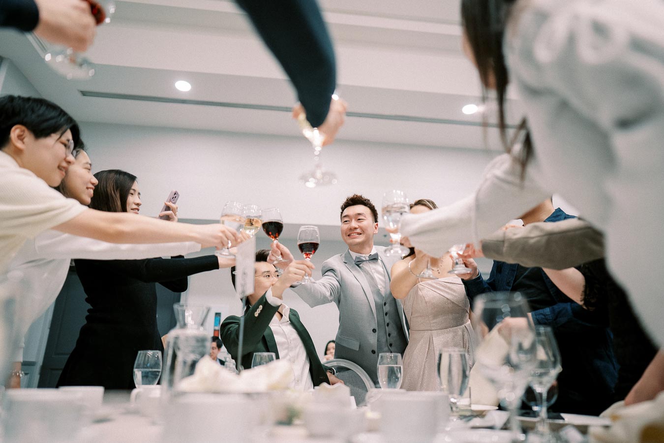 A group of people raising glasses in a celebratory toast at a formal event, featuring a man in a light gray suit smiling at the center of the gathering.