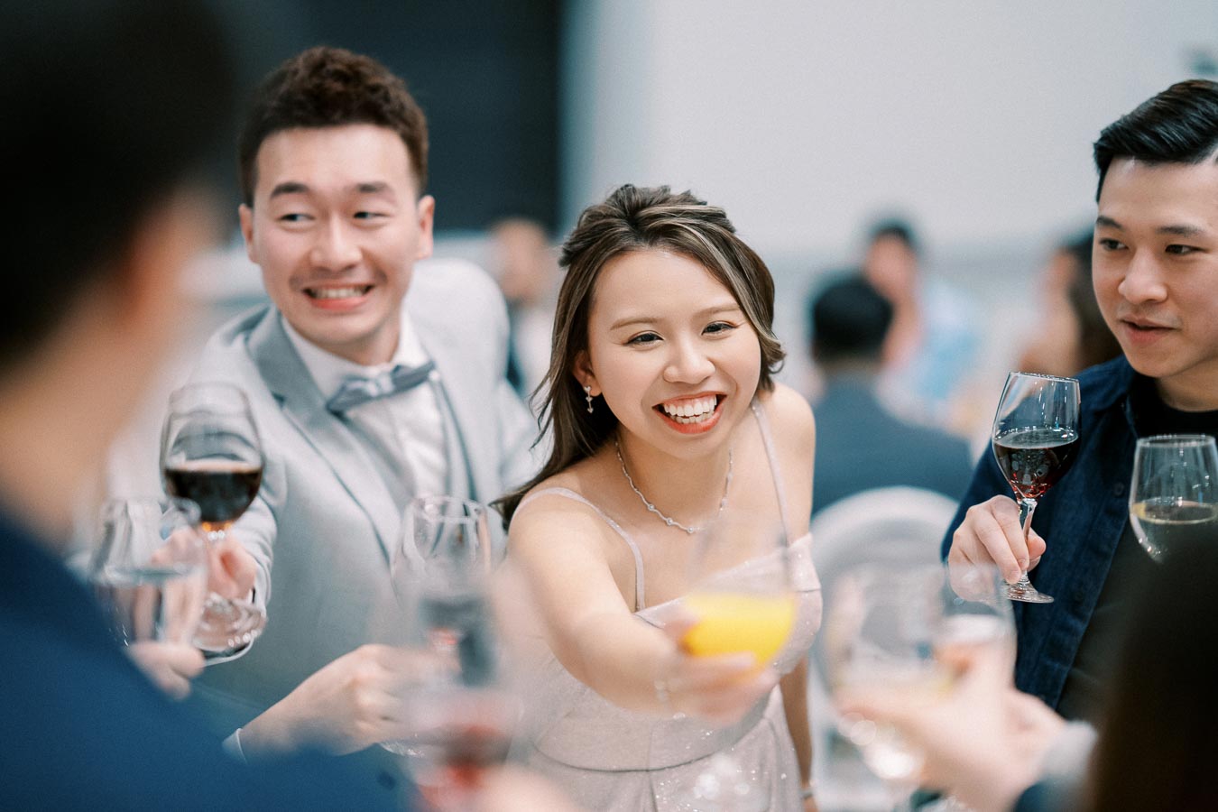 A group of people celebrating and clinking glasses at a social event, with a woman in a white dress smiling in the center, surrounded by friends holding various drinks.
