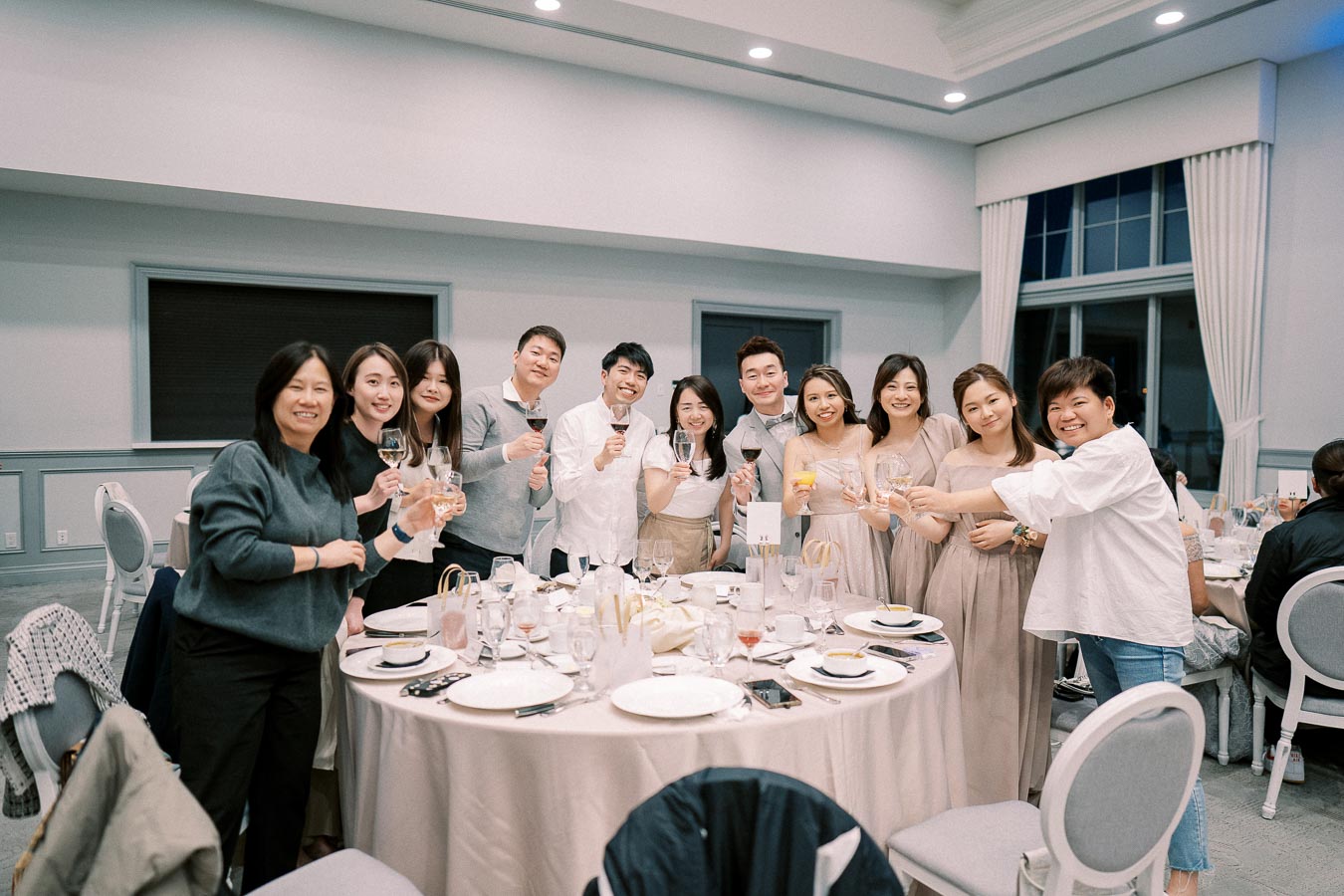 A group of people celebrating at an indoor party, smiling and holding glasses around a decorated table with white plates and silverware.