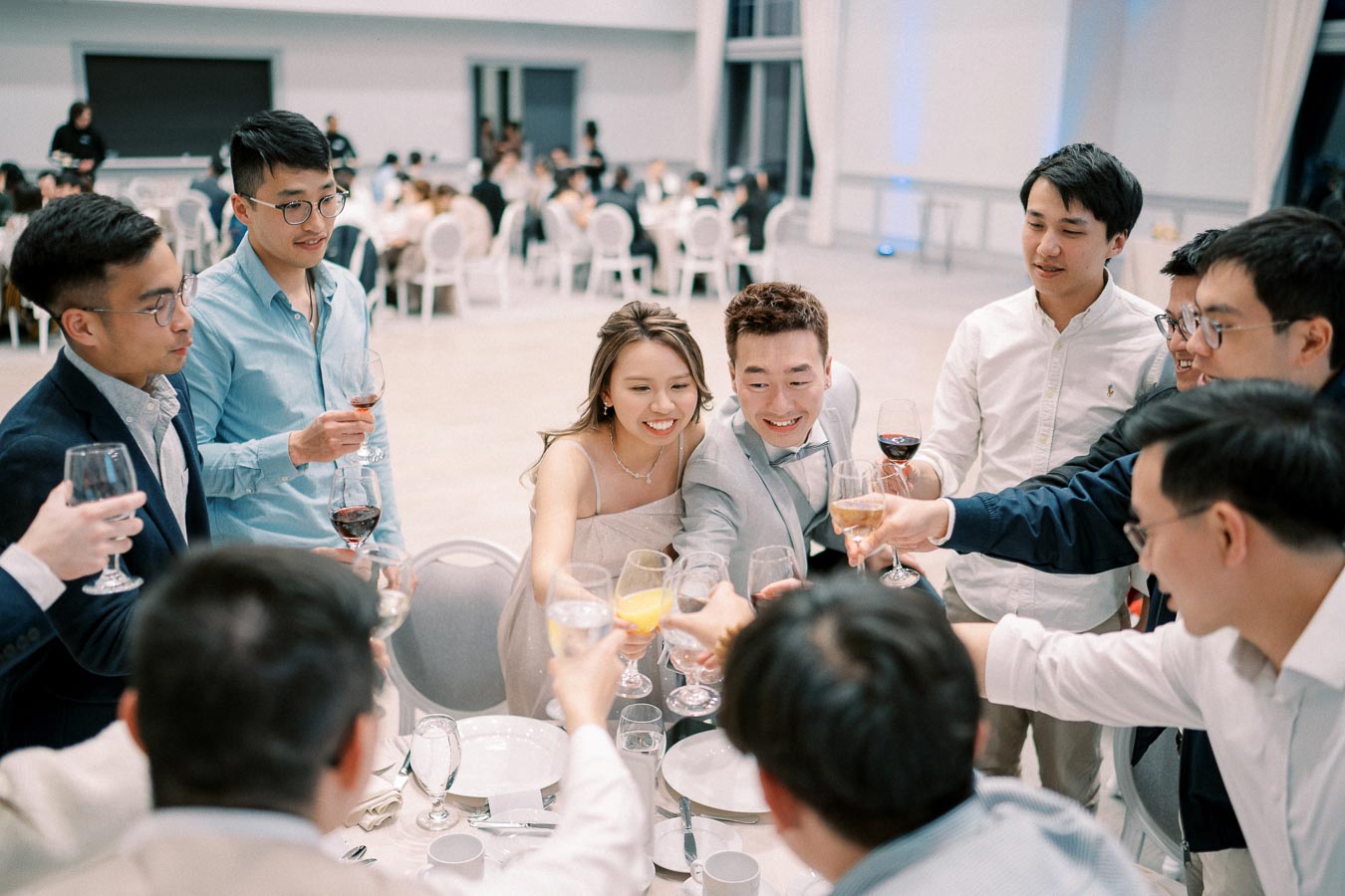 A group of elegantly dressed people enjoying a celebratory toast with glasses of wine at a formal event inside a spacious banquet hall.