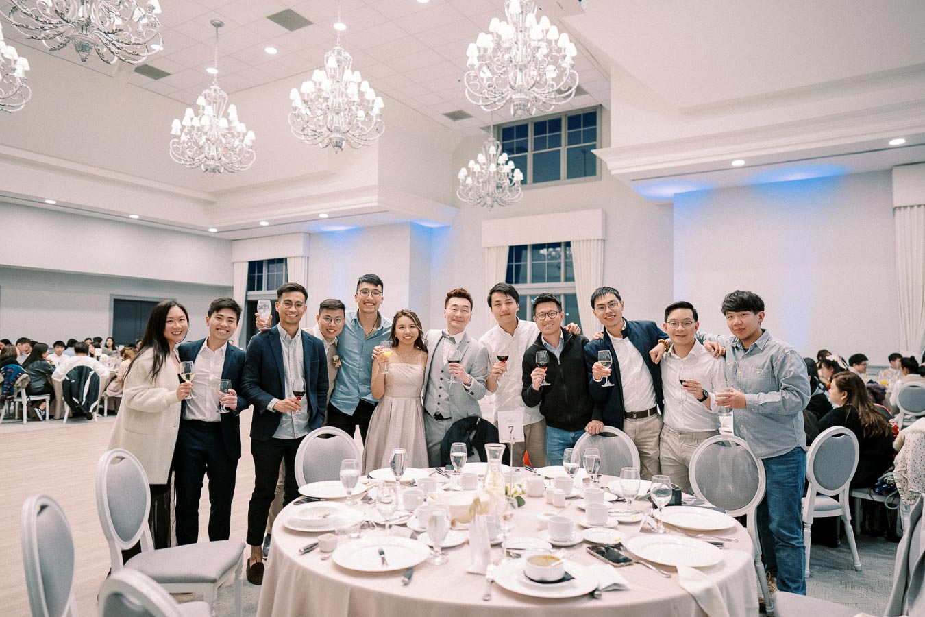 A group of people enjoying a formal event in a banquet hall with elegant chandeliers, toasting with drinks around a beautifully set dining table.