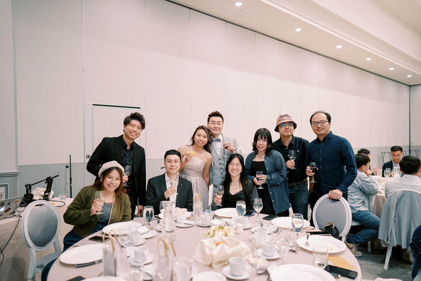 A group of people smiling and raising glasses at a formal event, seated around a table set with plates and utensils in a well-lit banquet hall.