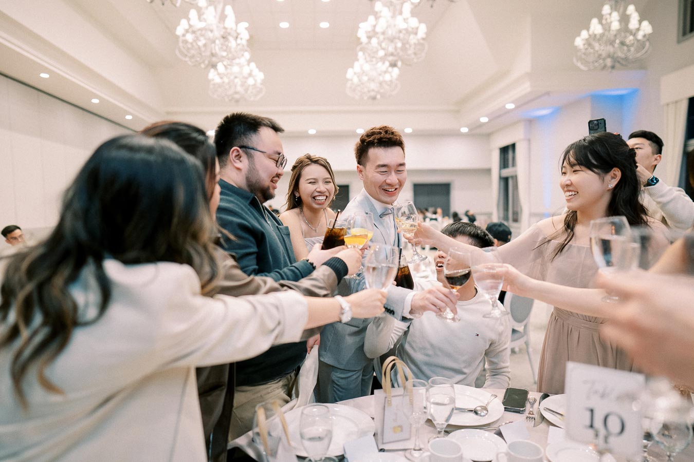 Group of people celebrating and clinking glasses at a wedding reception, with elegant chandeliers and a beautifully set table in the background.