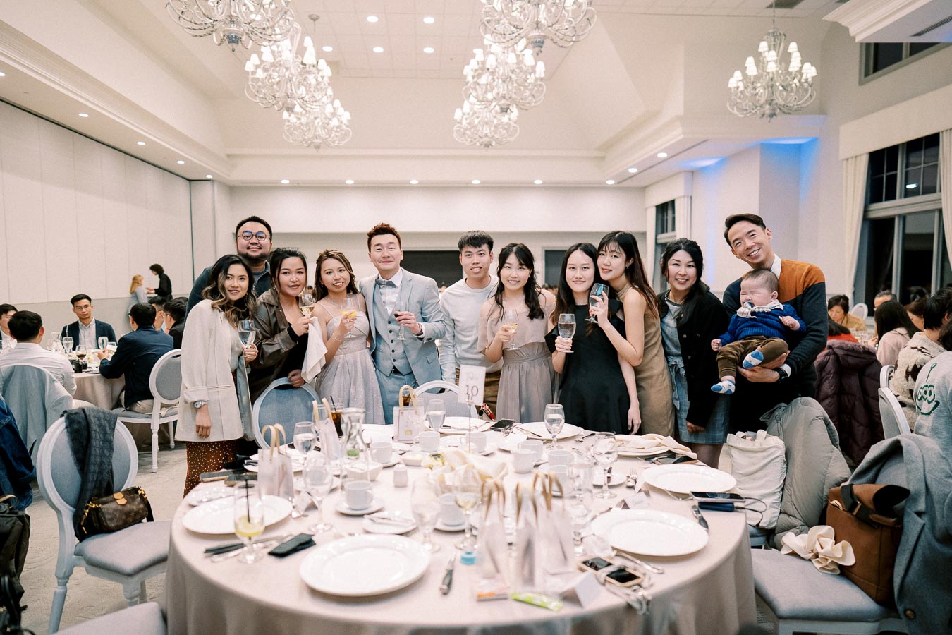 A cheerful group of people posing together at a formal event, holding drinks and smiling at the camera. The elegantly set table in the foreground indicates a celebratory occasion in a beautifully lit banquet hall.