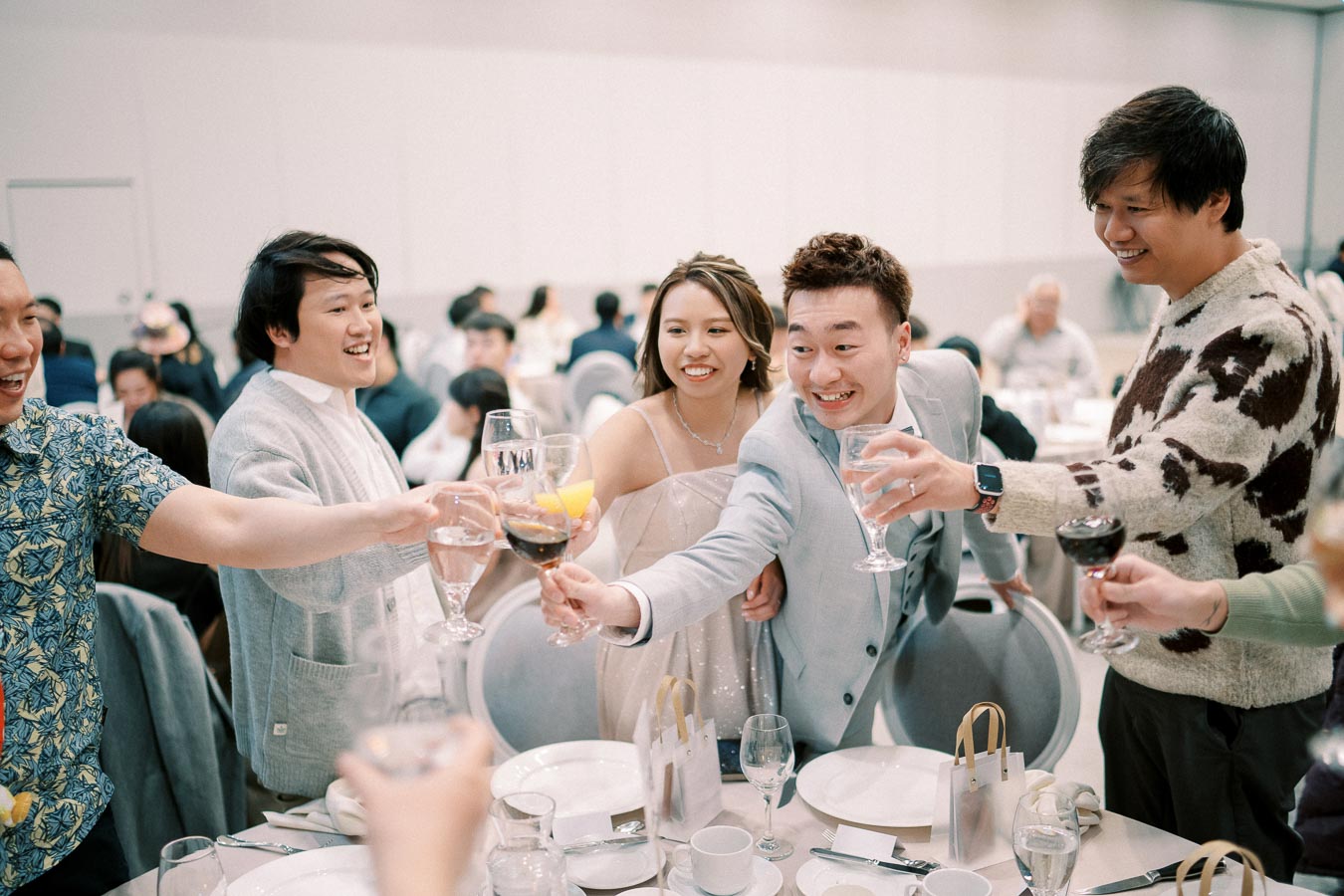 A group of people celebrating with a toast at a festive gathering, dressed in formal and semi-formal attire, and smiling around a table set with plates, glasses, and gift bags.