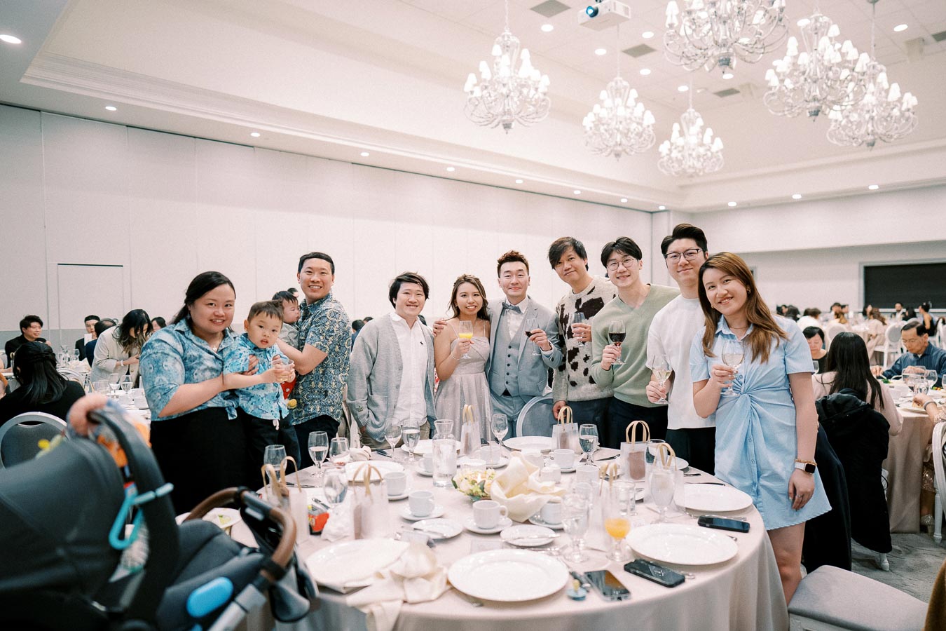 A group of people celebrating at a banquet, holding drinks and smiling around a table set for a formal event, with elegant chandeliers above and guests in the background.