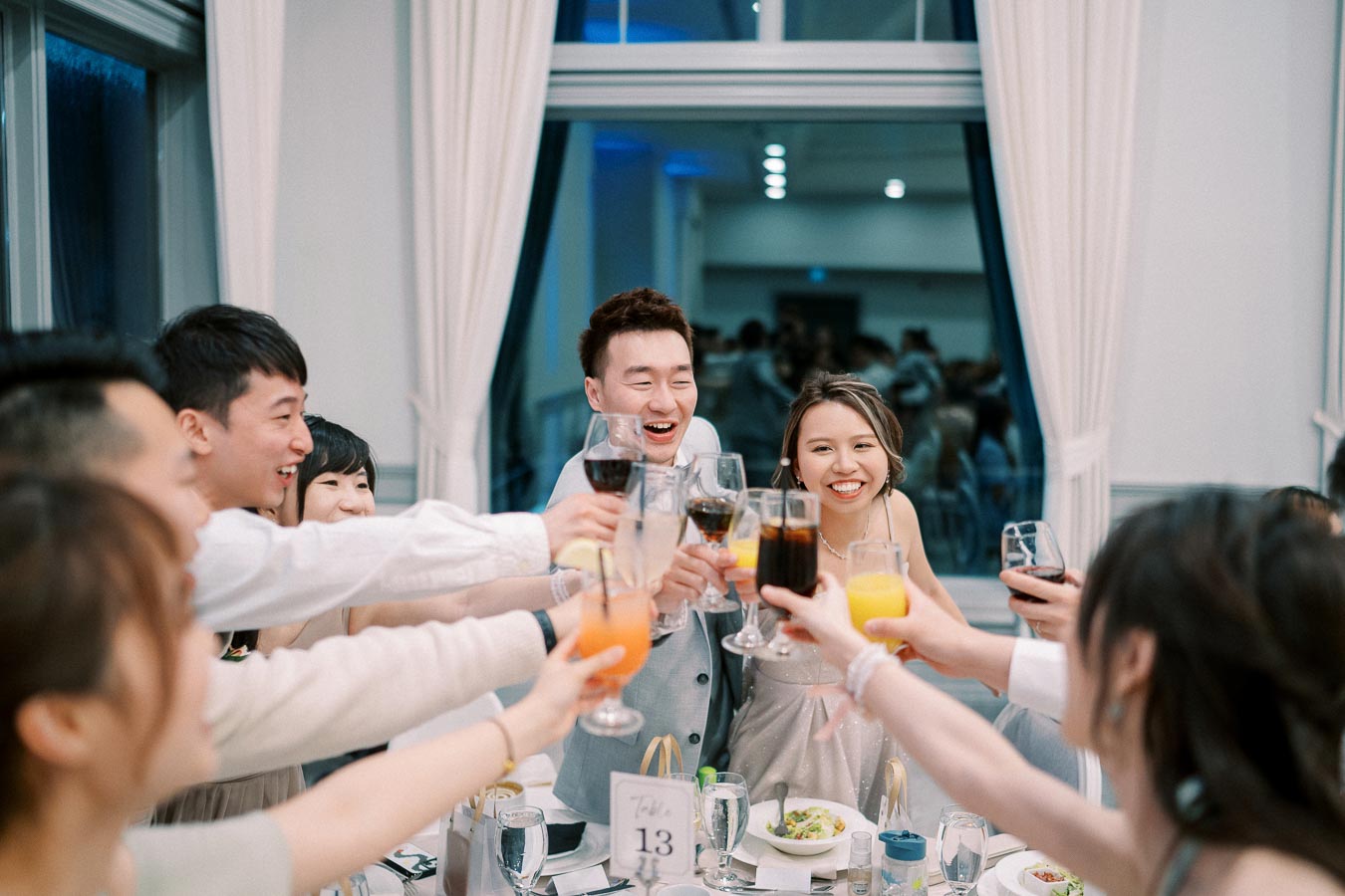 A group of friends raising glasses in a celebratory toast at a wedding reception, with a happy bride and groom at the center, surrounded by a festive atmosphere, table number 13 visible.