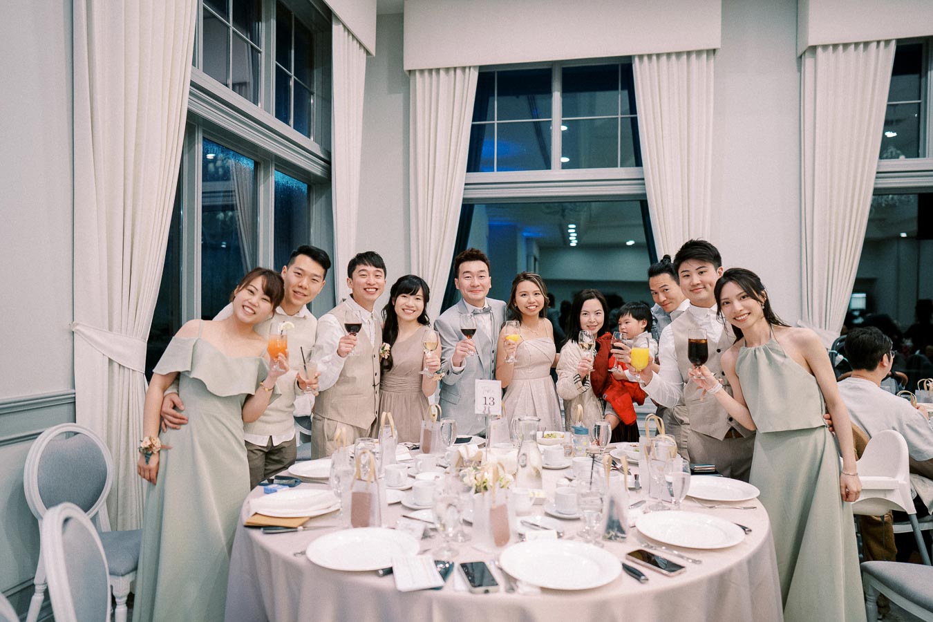 Guests celebrating at a formal event, dressed in elegant attire and holding drinks, gathered around a beautifully set banquet table with large windows in the background.