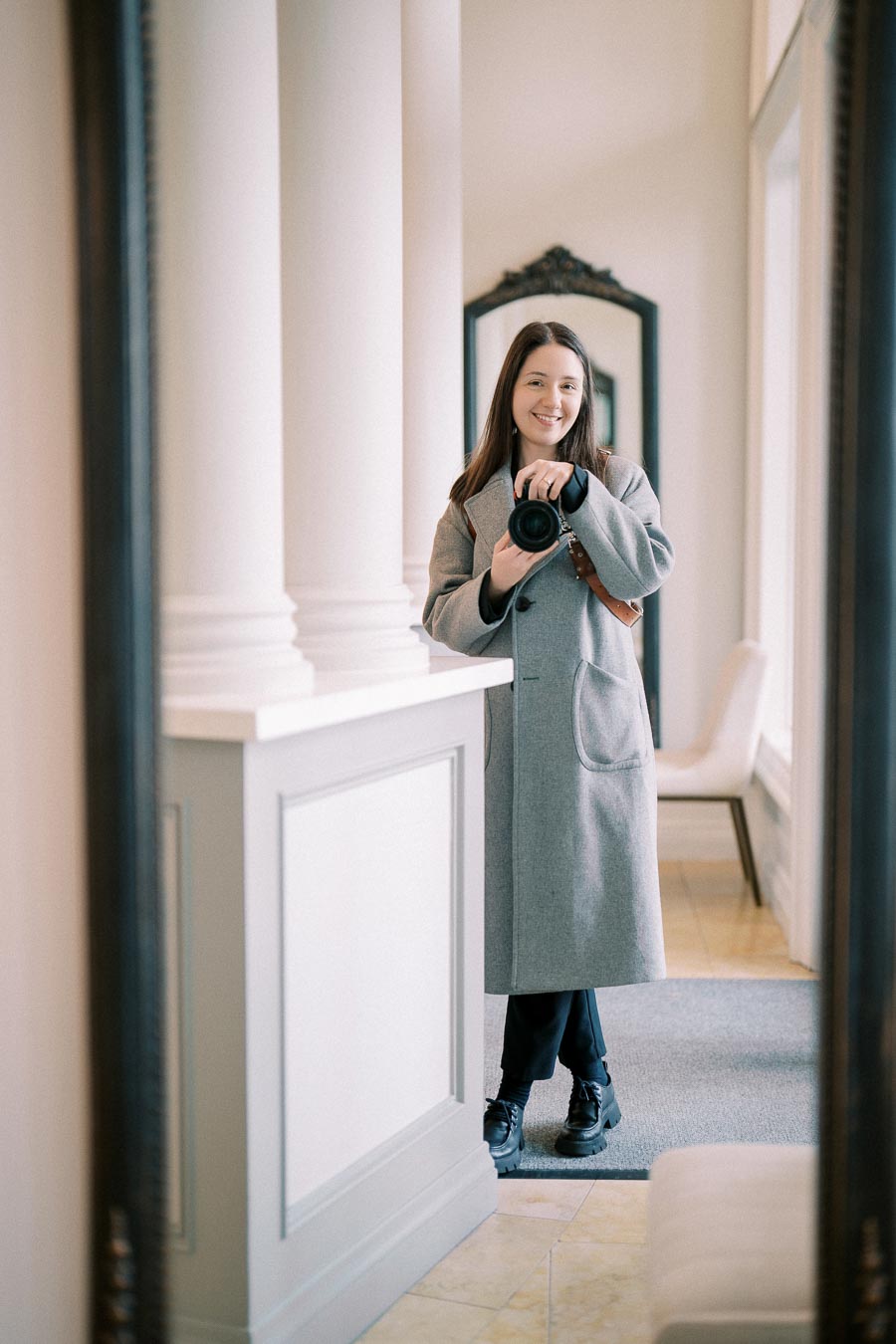 A woman in a gray coat stands in a luxurious hallway, capturing her reflection in a mirror with a camera.