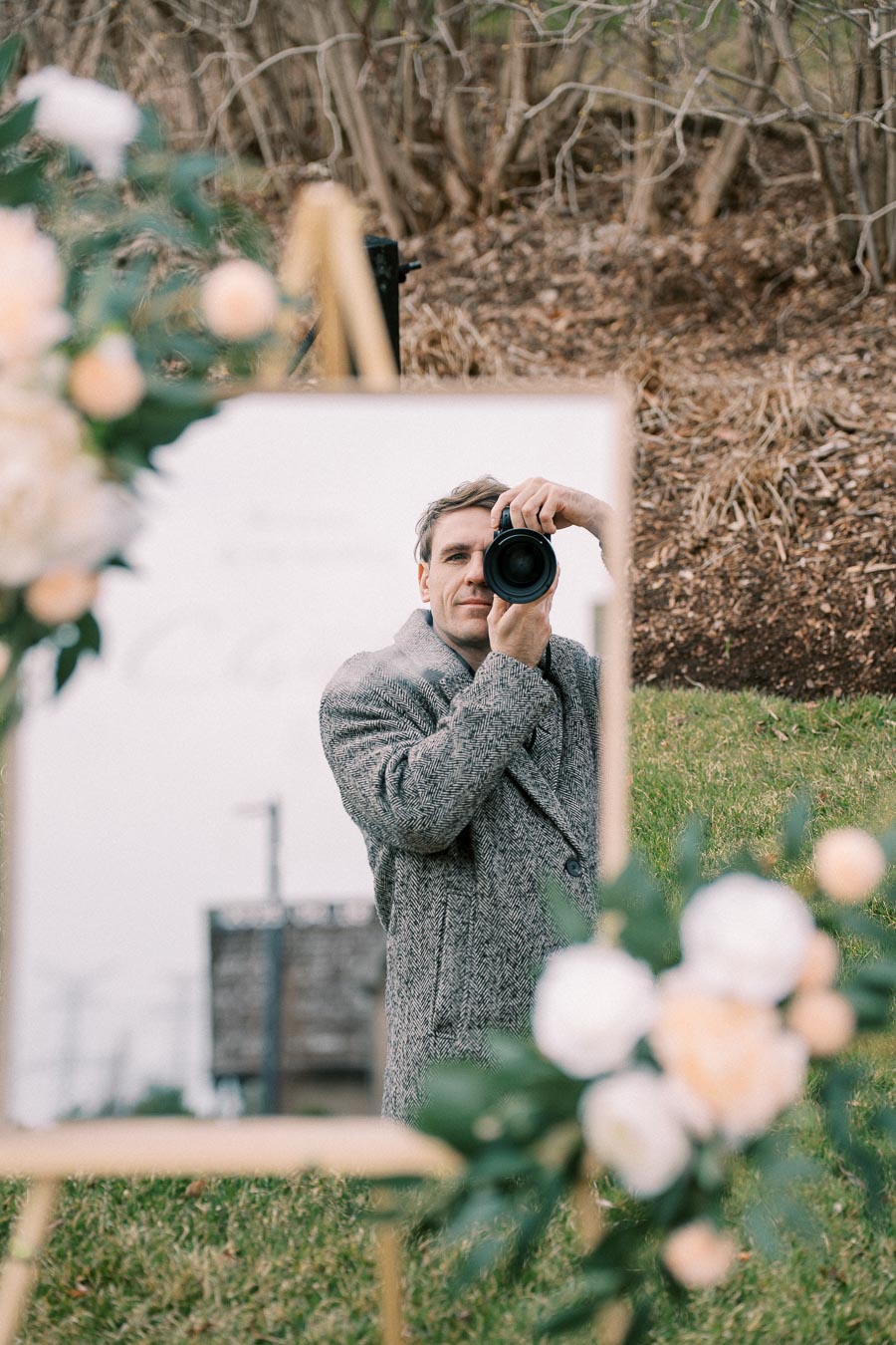 Photographer capturing self-portrait in a mirror with floral decorations in an outdoor setting, wearing a gray coat.