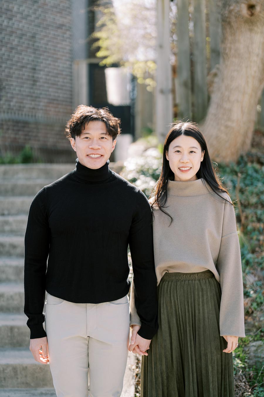 A couple smiling and holding hands while standing on outdoor steps, surrounded by greenery.
