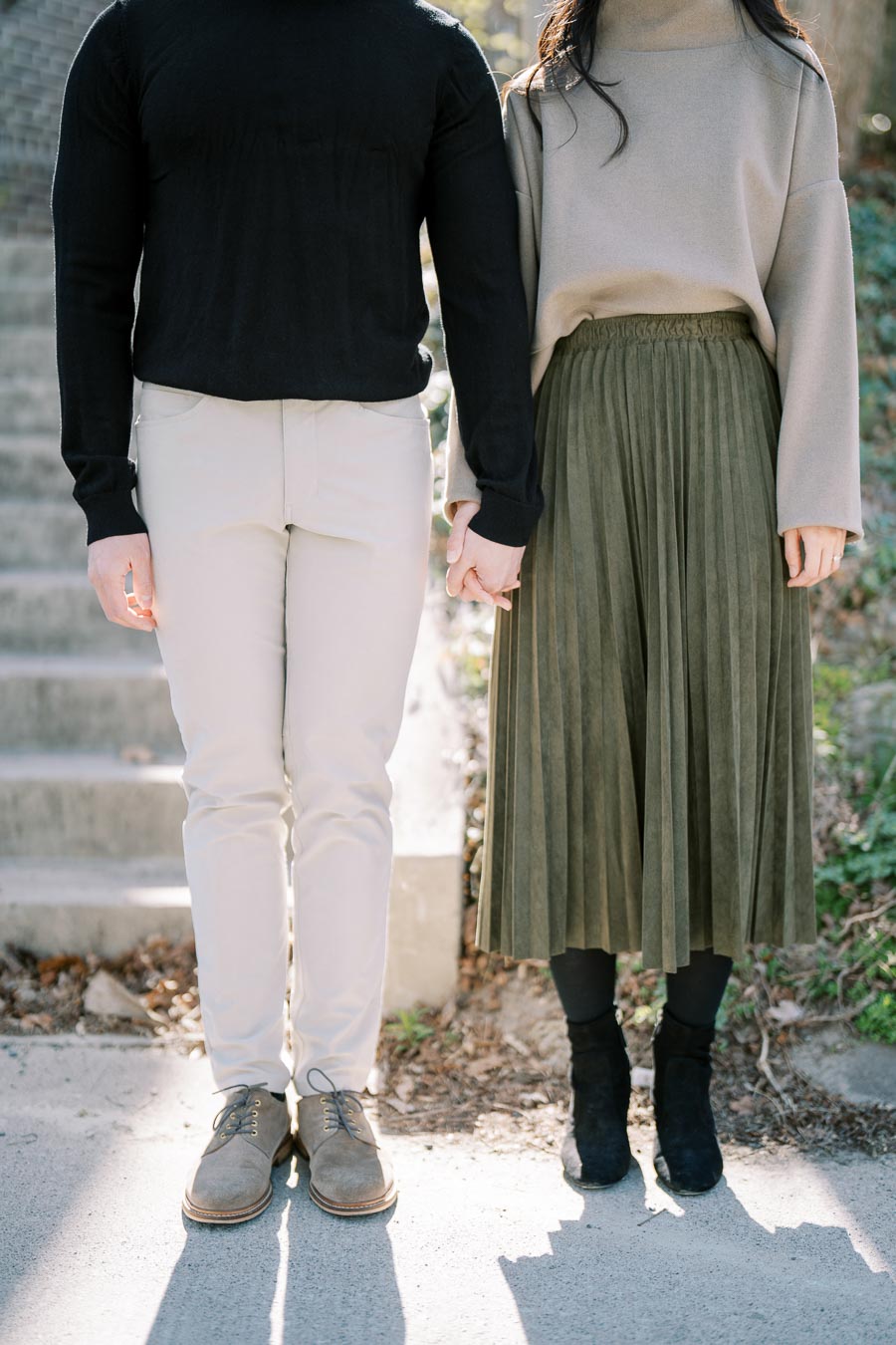 A couple holding hands, dressed in casual attire, standing on a concrete path with steps in the background.