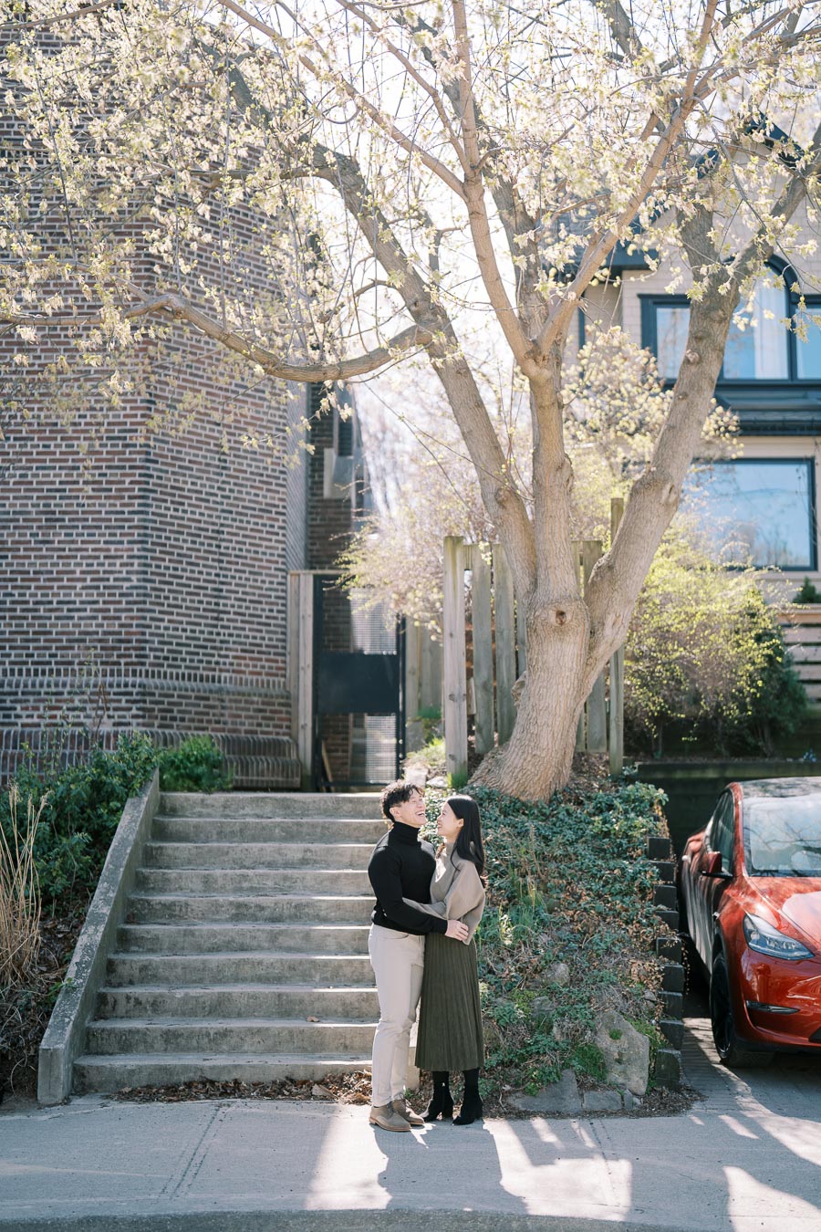 A couple shares a joyful moment under a blooming tree beside a brick building, with a red car parked nearby.