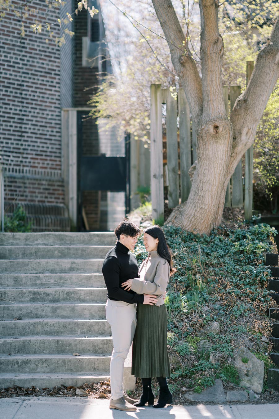 A couple shares a tender moment on a sunny day, standing in front of a staircase with lush greenery and a large tree nearby.