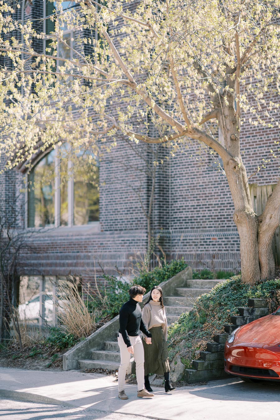 A couple enjoys a sunny day, walking hand in hand past a brick building and a tree with budding leaves.