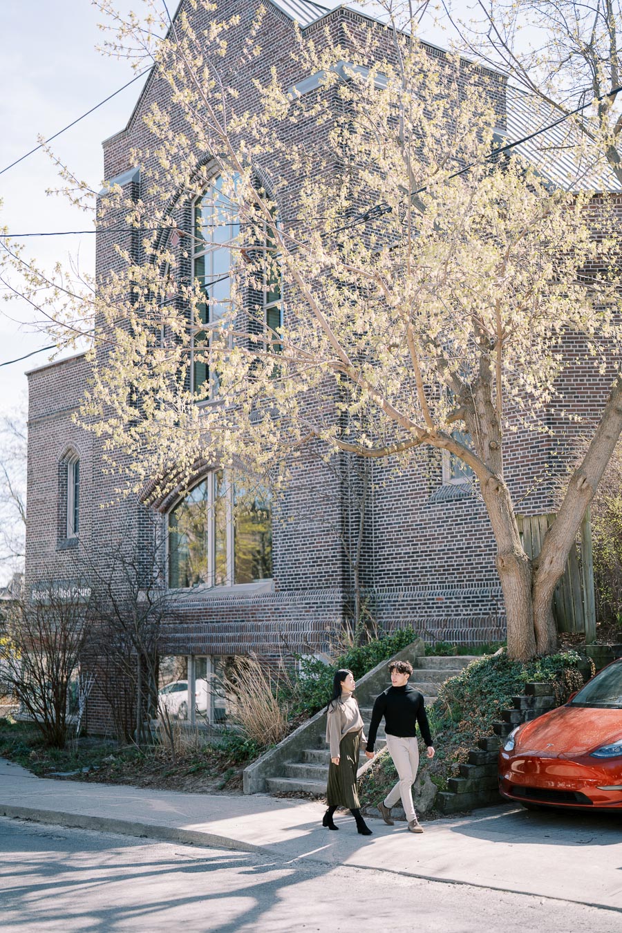 A couple walks past a brick building with blooming trees, enjoying a sunny day.