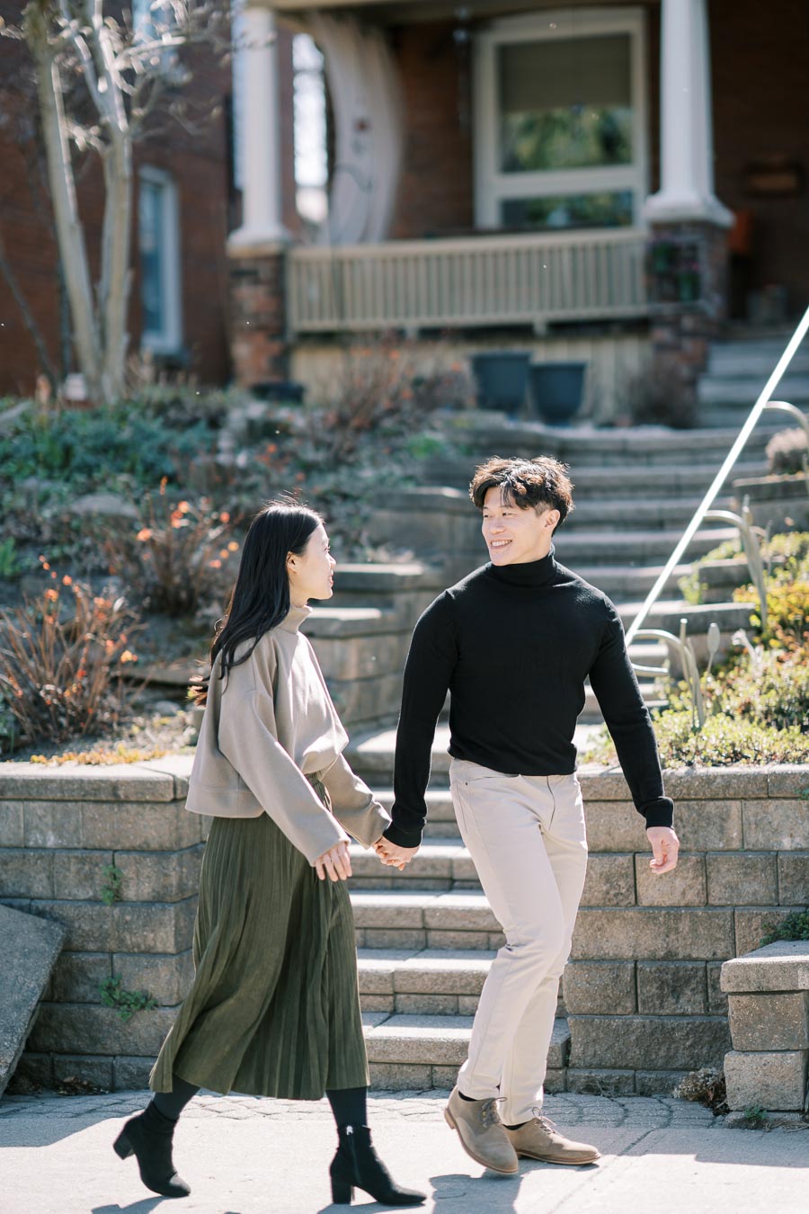 A couple enjoys a sunny day, holding hands as they walk past a brick house with steps and garden plants.