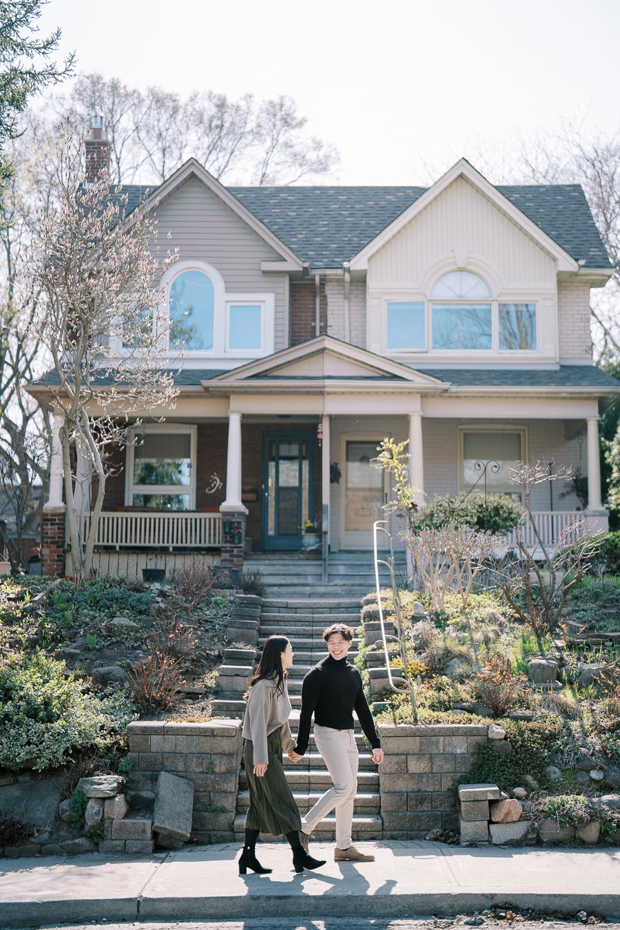 A couple walks hand in hand in front of a charming two-story house with a well-maintained garden.