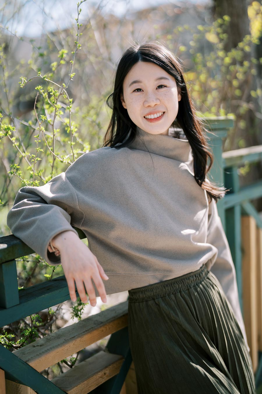 A woman enjoying a sunny day outdoors, leaning on a wooden railing surrounded by greenery.