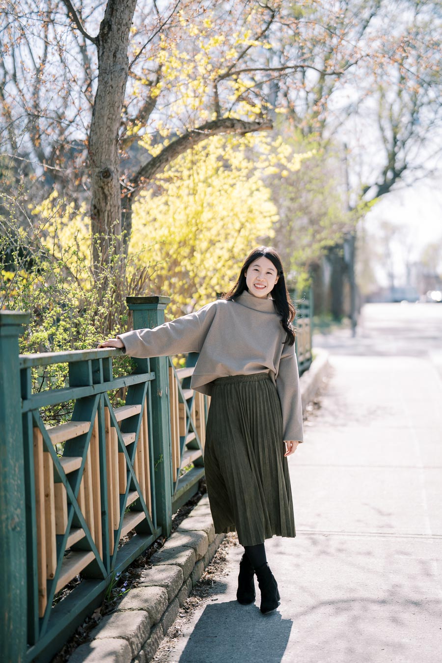 A woman smiles while standing on a sidewalk next to a decorative fence, surrounded by spring foliage.
