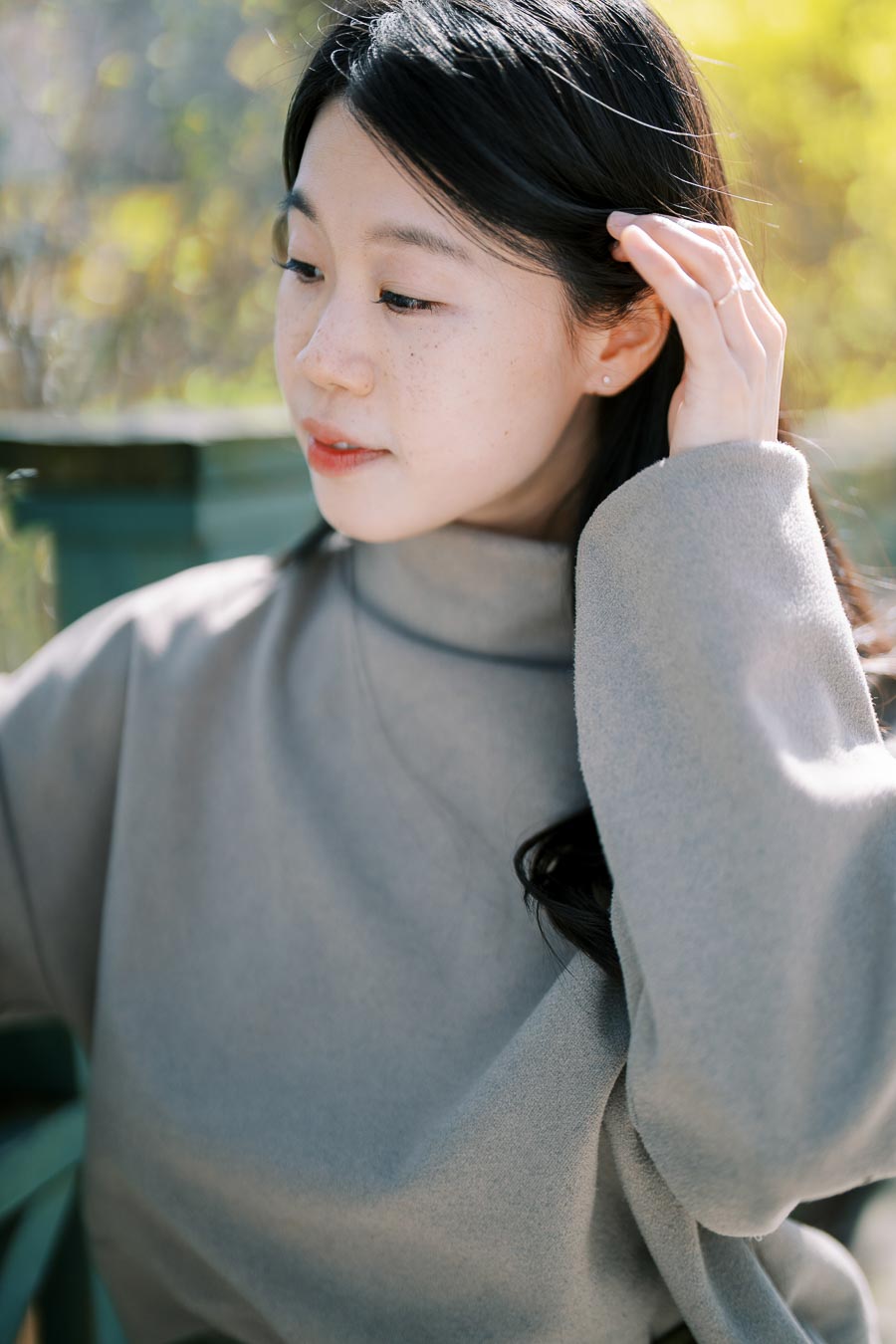 A woman in a gray turtleneck sweater gently touches her hair while looking down, set against a soft, blurred green background.