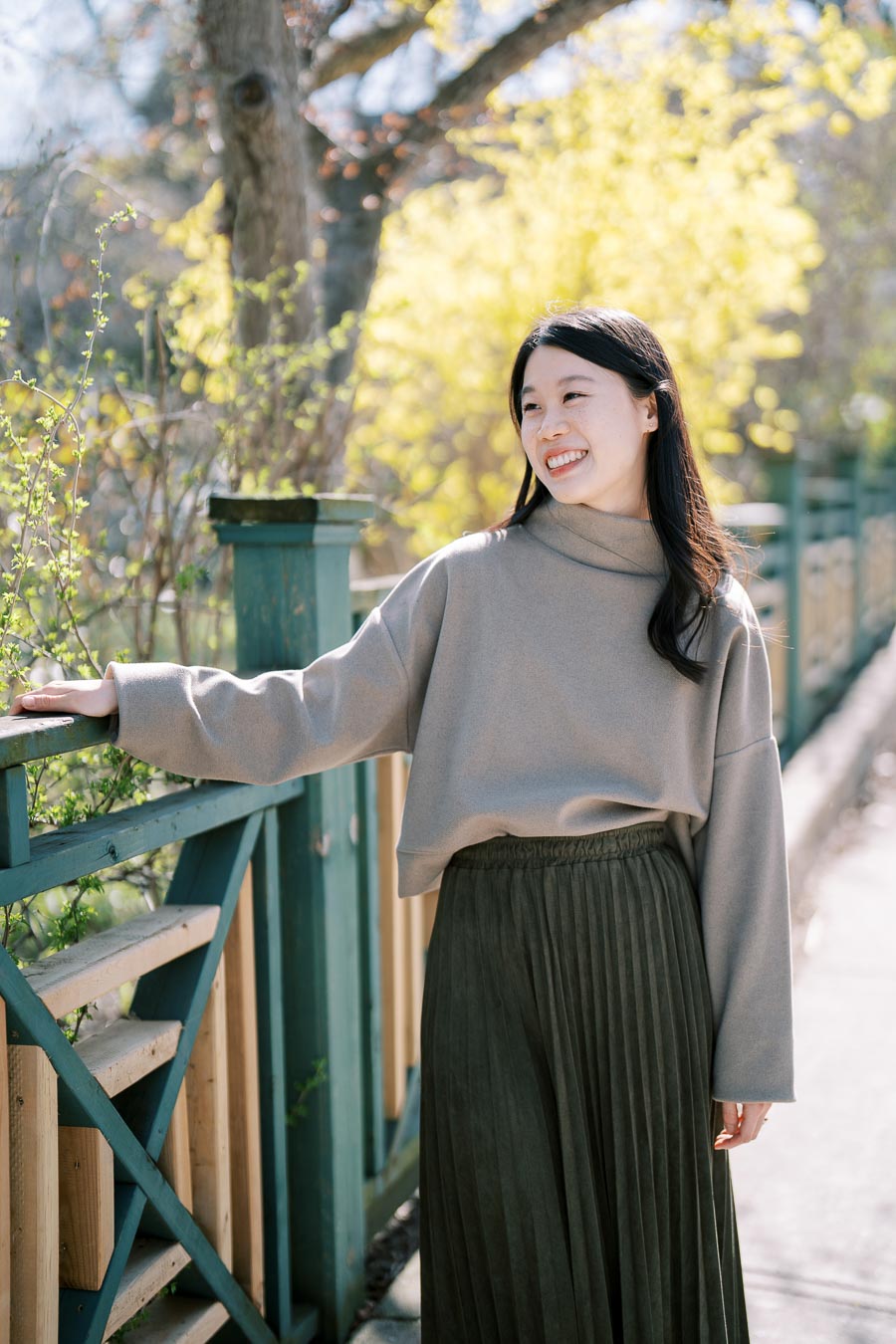 A woman enjoys a sunny day outdoors, leaning on a wooden fence surrounded by spring foliage.