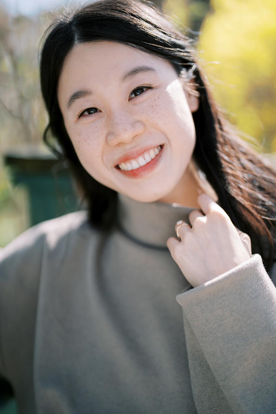 A woman smiling warmly in an outdoor setting, with a soft focus on the greenery behind her.