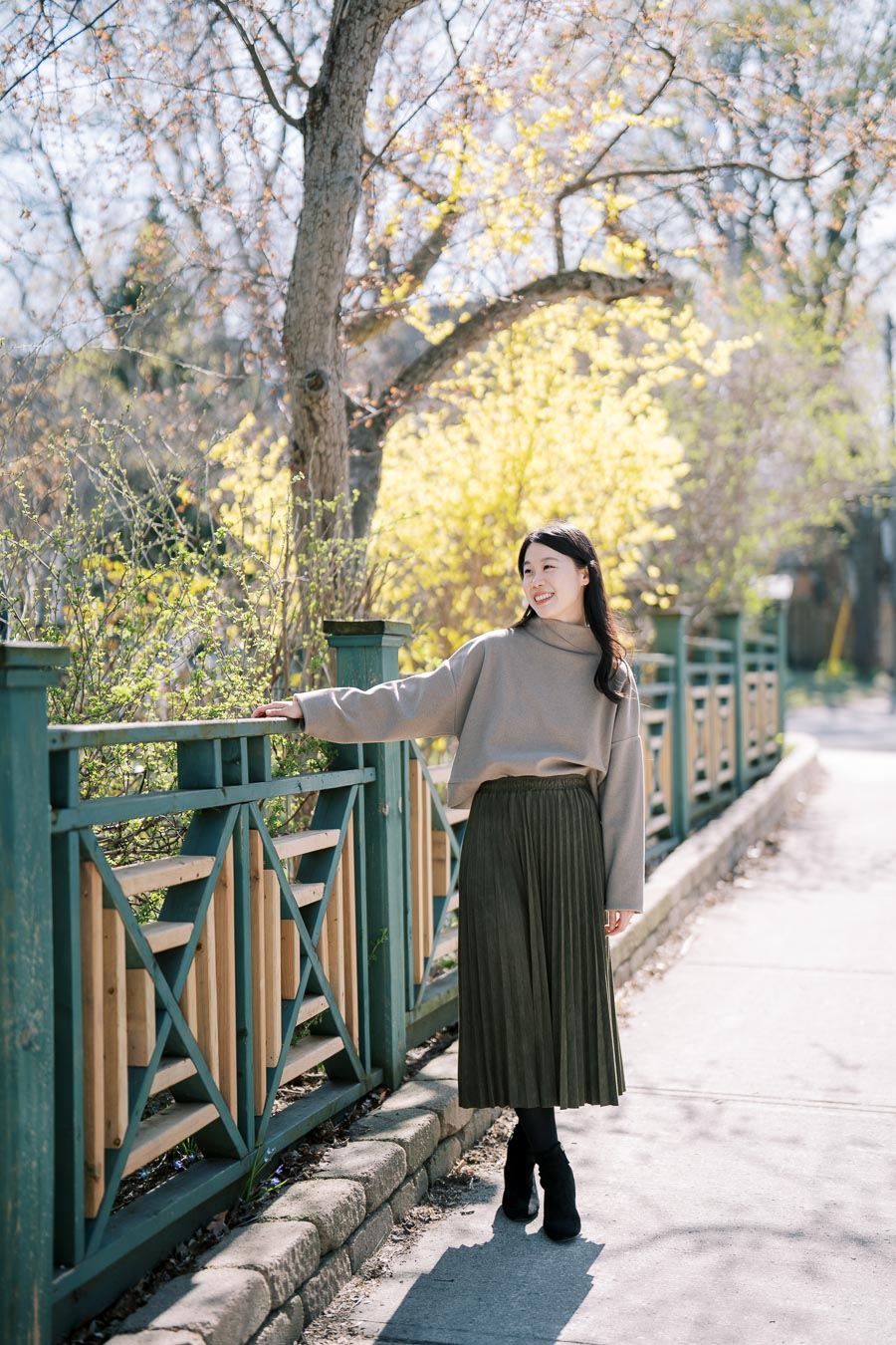 A woman enjoys a sunny spring day, standing by a decorative wooden fence with blooming trees in the background.