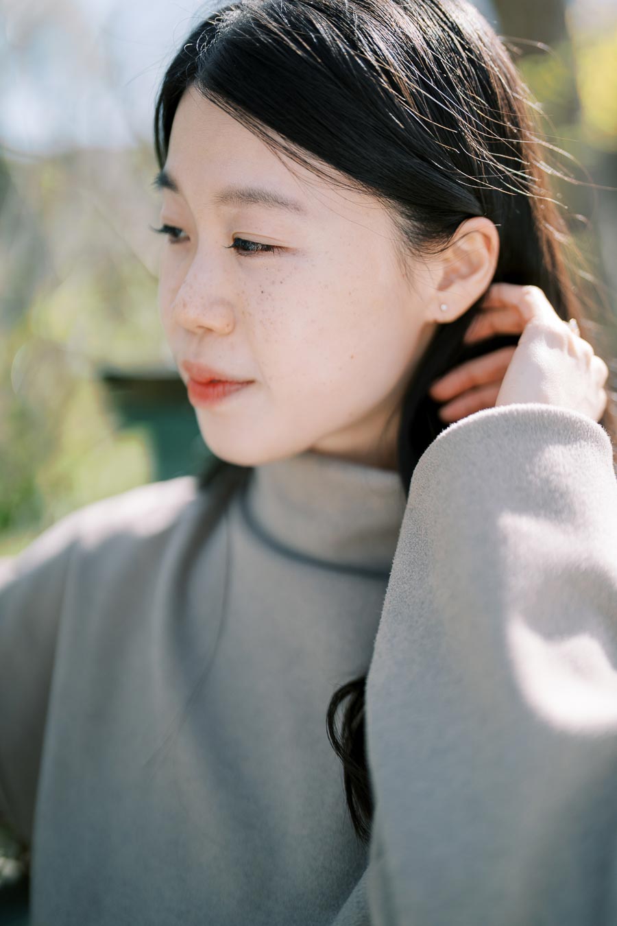 A woman in a gray turtleneck sweater gazes thoughtfully to the side in a serene outdoor setting.