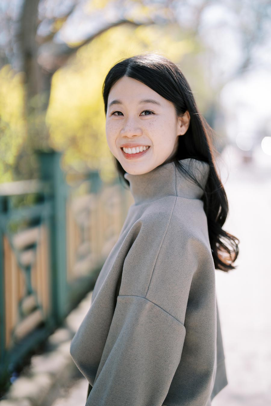 A woman smiling outdoors, wearing a gray coat, with a blurred background of trees and a fence.