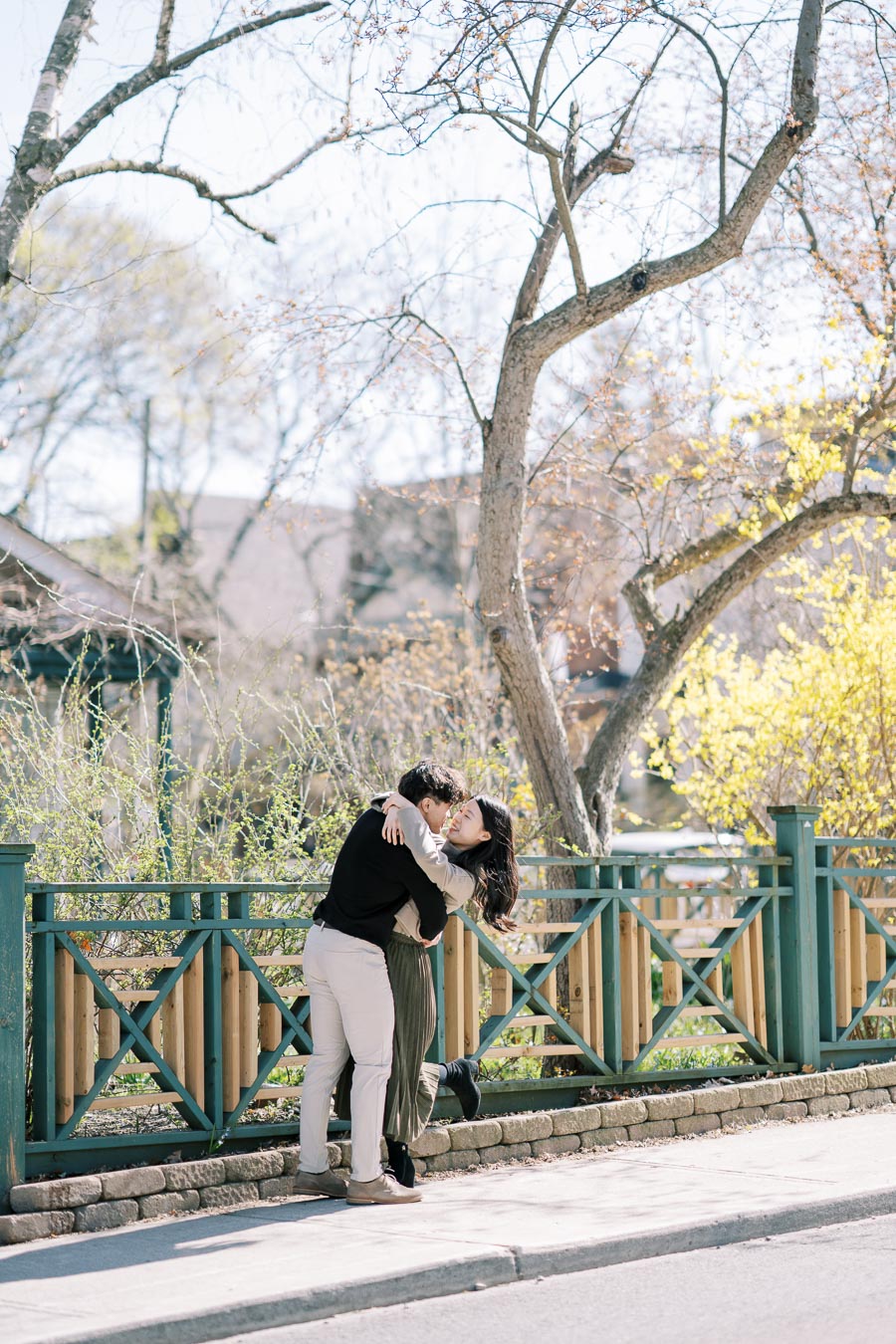 A joyful couple shares a loving embrace on a sunny day near a wooden fence, surrounded by budding trees.